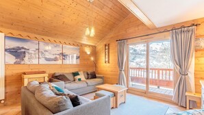 A cozy wooden cabin interior with a sofa, a small table, and a view of snowy mountains through large windows. - 50m from ski slopes, 3rd floor, view ski slopes, fitness, swimming pool, sauna, hammam, balcony, tv (Saint-Martin-de-Belleville)