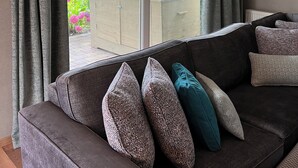 Un salon moderne avec un canapé gris foncé, de nombreux coussins à motifs et une vue sur un jardin grâce à de grandes fenêtres.