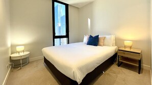 A bedroom with a large bed, two bedside tables with lamps, a window with a view of buildings, and a carpeted floor.