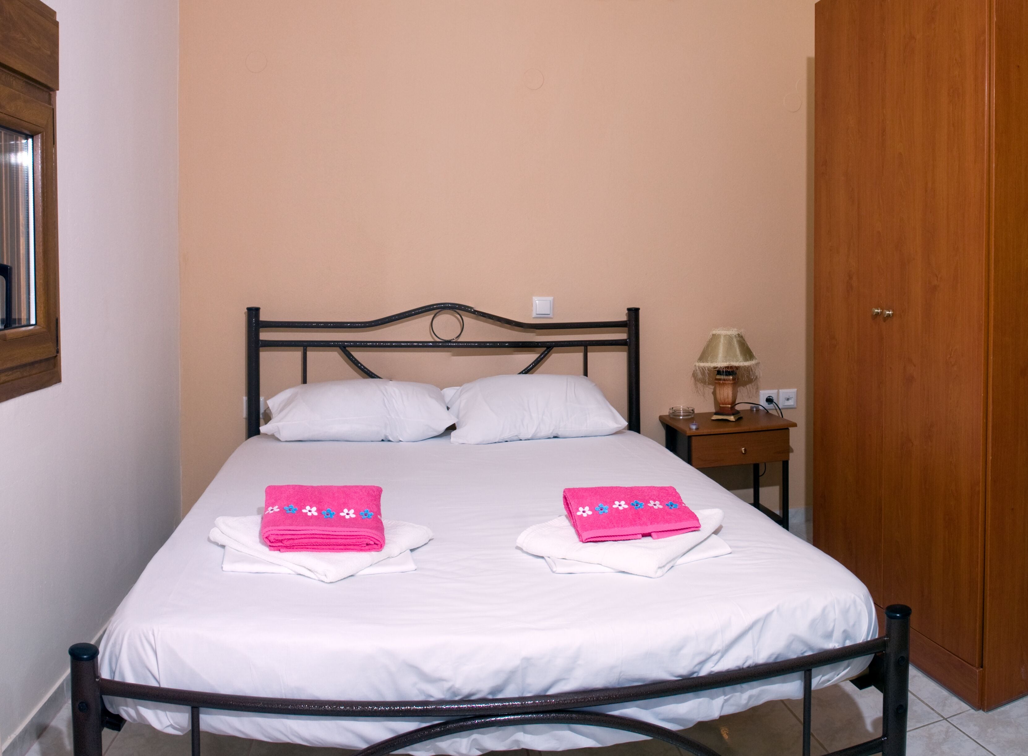 A single bed with white bedding, two pink towels with floral designs, a bedside lamp, and a wooden wardrobe.