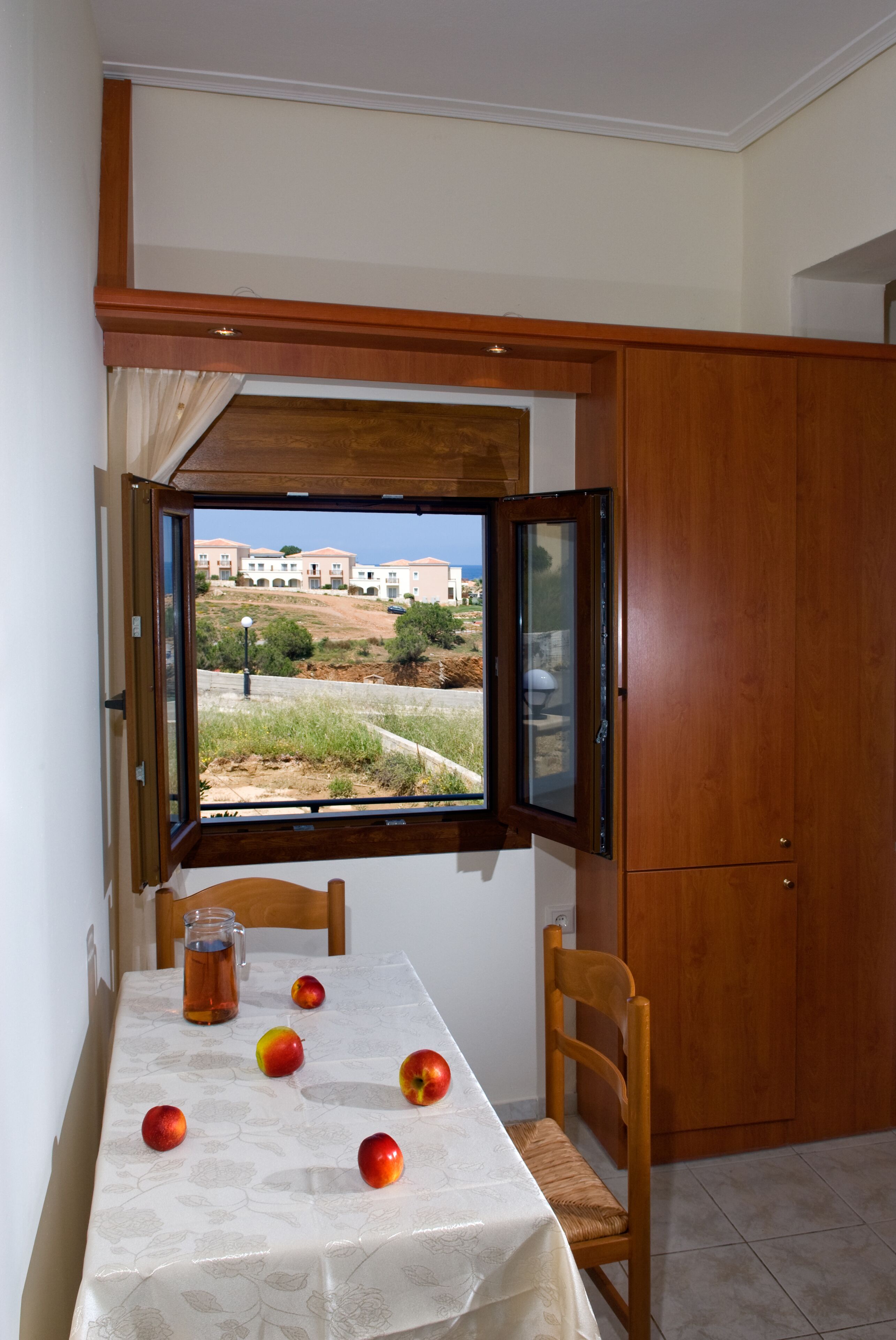 A dining area with a table set for two, a window with an open view of buildings and greenery, and a wooden cabinet to the right.