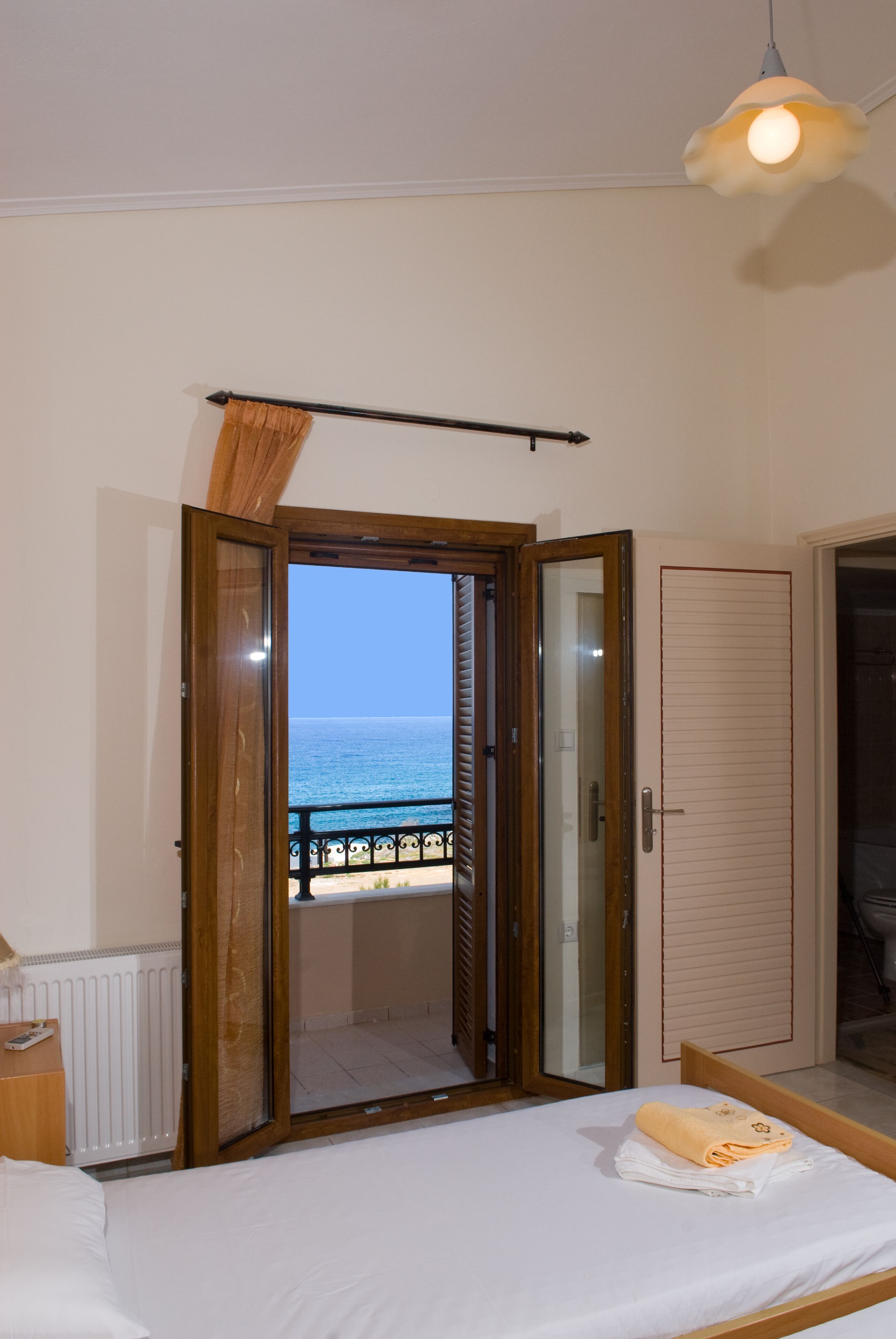 A bedroom with a bed, a wooden headboard, a nightstand, a towel, a wardrobe, and a view of the sea through an open door.