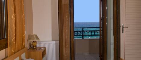 A bedroom with a bed, a nightstand with a lamp, a window with curtains, and a view of the sea through an open door.