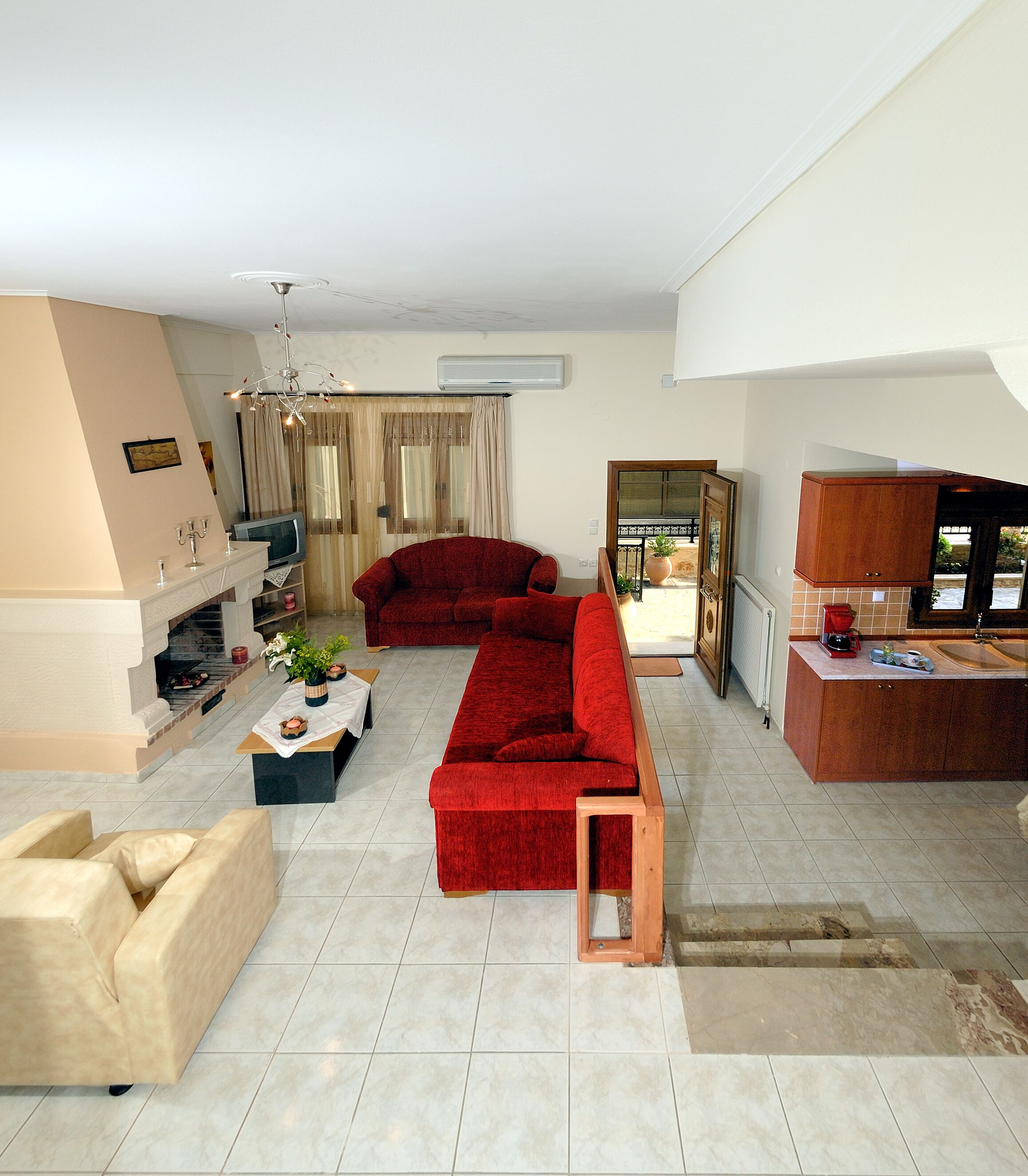 A spacious living room with a red sofa, a beige armchair, and a wooden coffee table. There is a kitchen area with wooden cabinets and a sink.