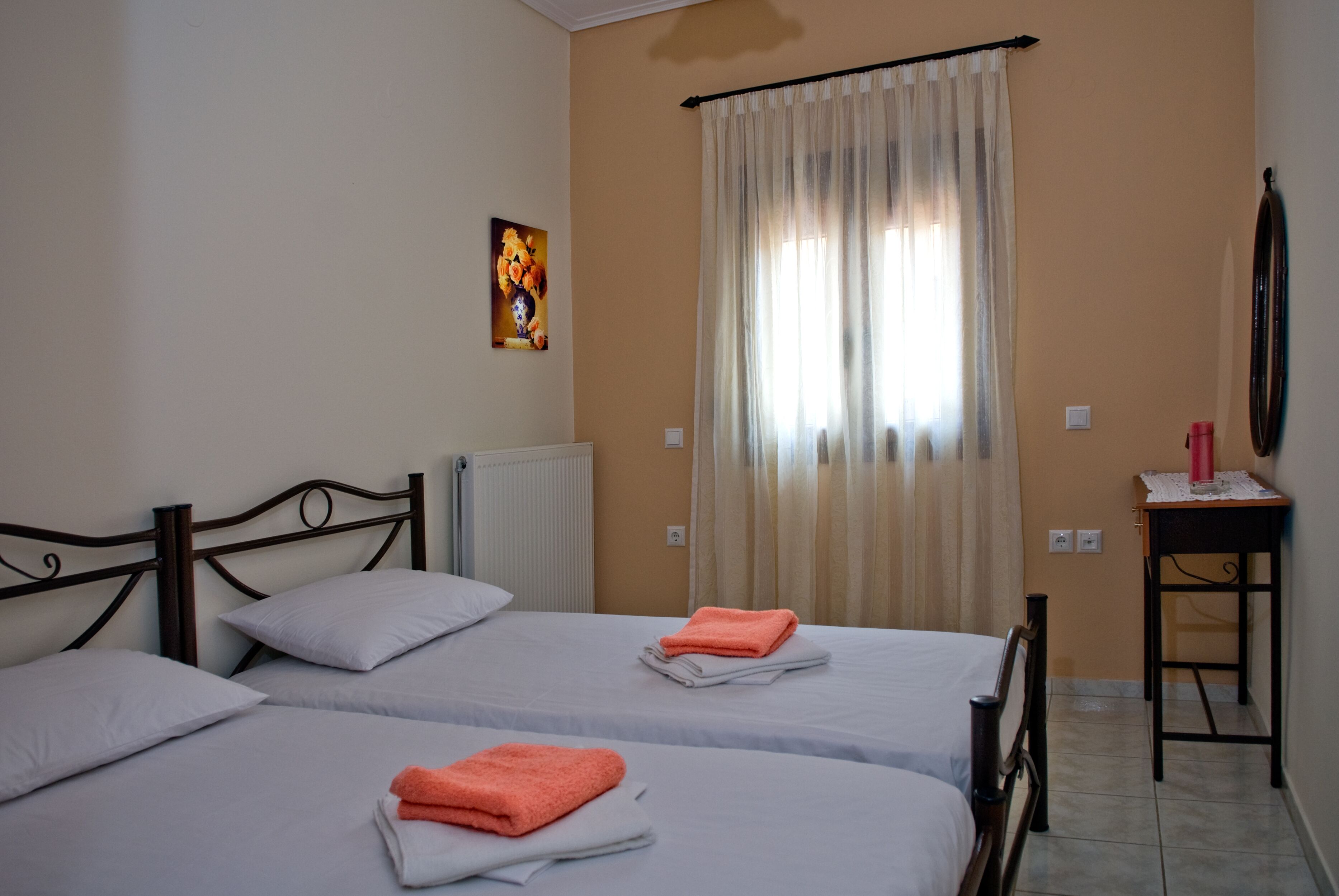 A room with two beds, a radiator, a painting on the wall, a table with a lamp, and a window with curtains.