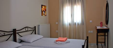 A room with two beds, a radiator, a painting on the wall, a table with a lamp, and a window with curtains.