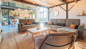 A spacious living area with two sofas, a coffee table, and a wicker chair. A staircase leads to an upper level with a kitchen and dining area. - Gîte un Air de Campagne (Gergny)