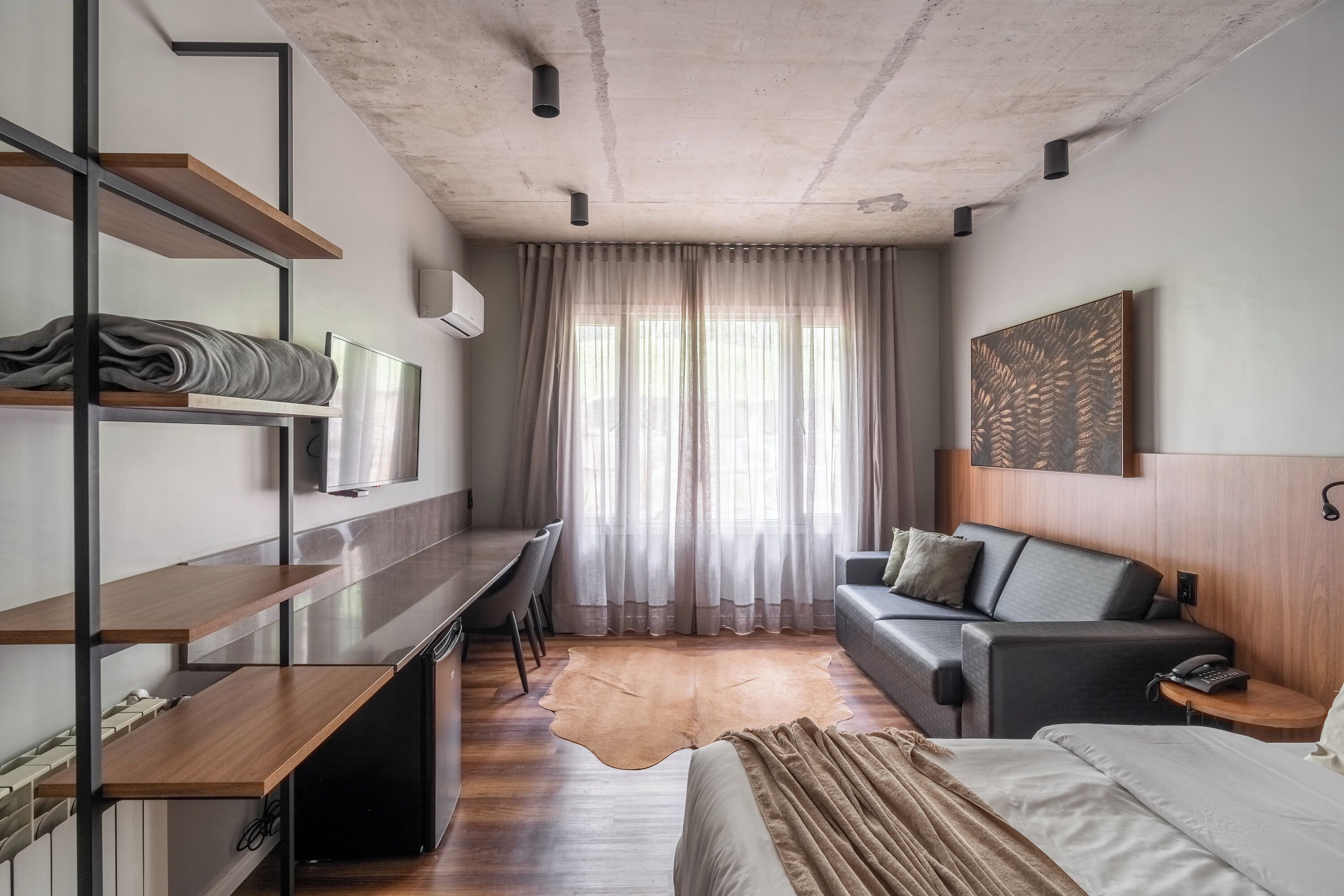 Una habitación de hotel moderna con cama, sofá, escritorio y un amplio ventanal con cortinas.