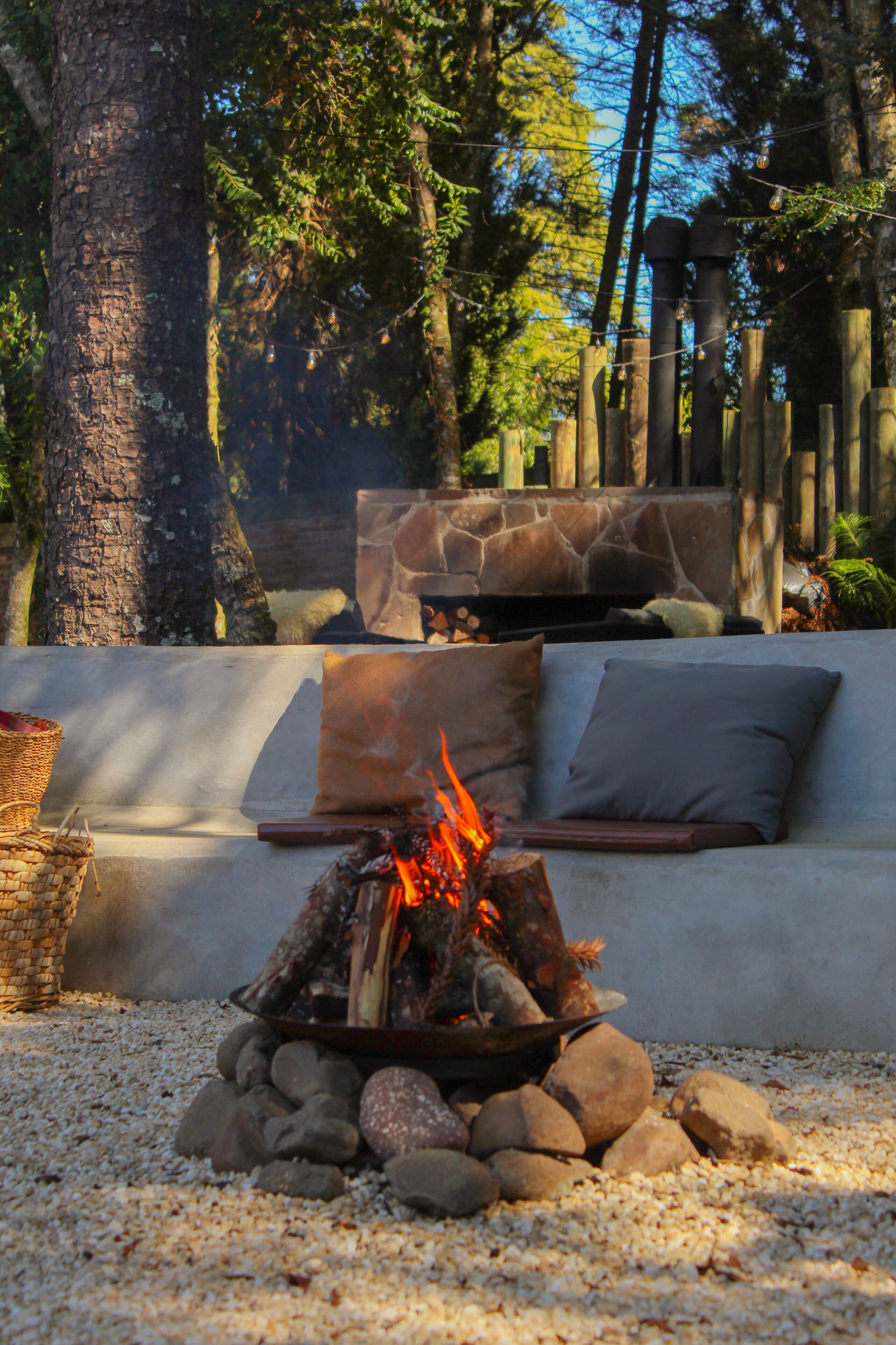 A fire pit with logs, surrounded by rocks, in a garden area with a stone bench and cushions.