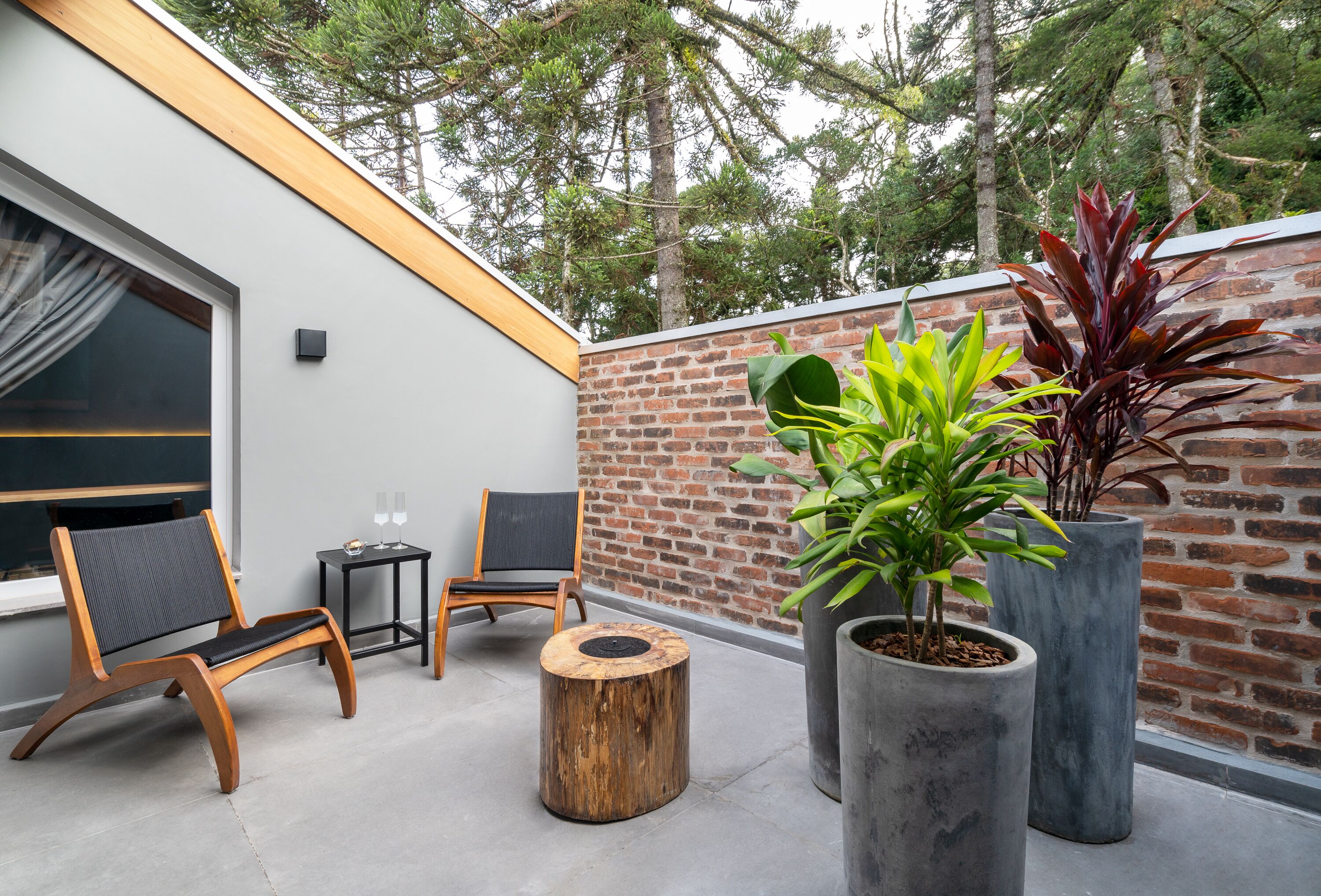 An outdoor seating area with two wooden chairs, a round wooden stool, and two large potted plants.