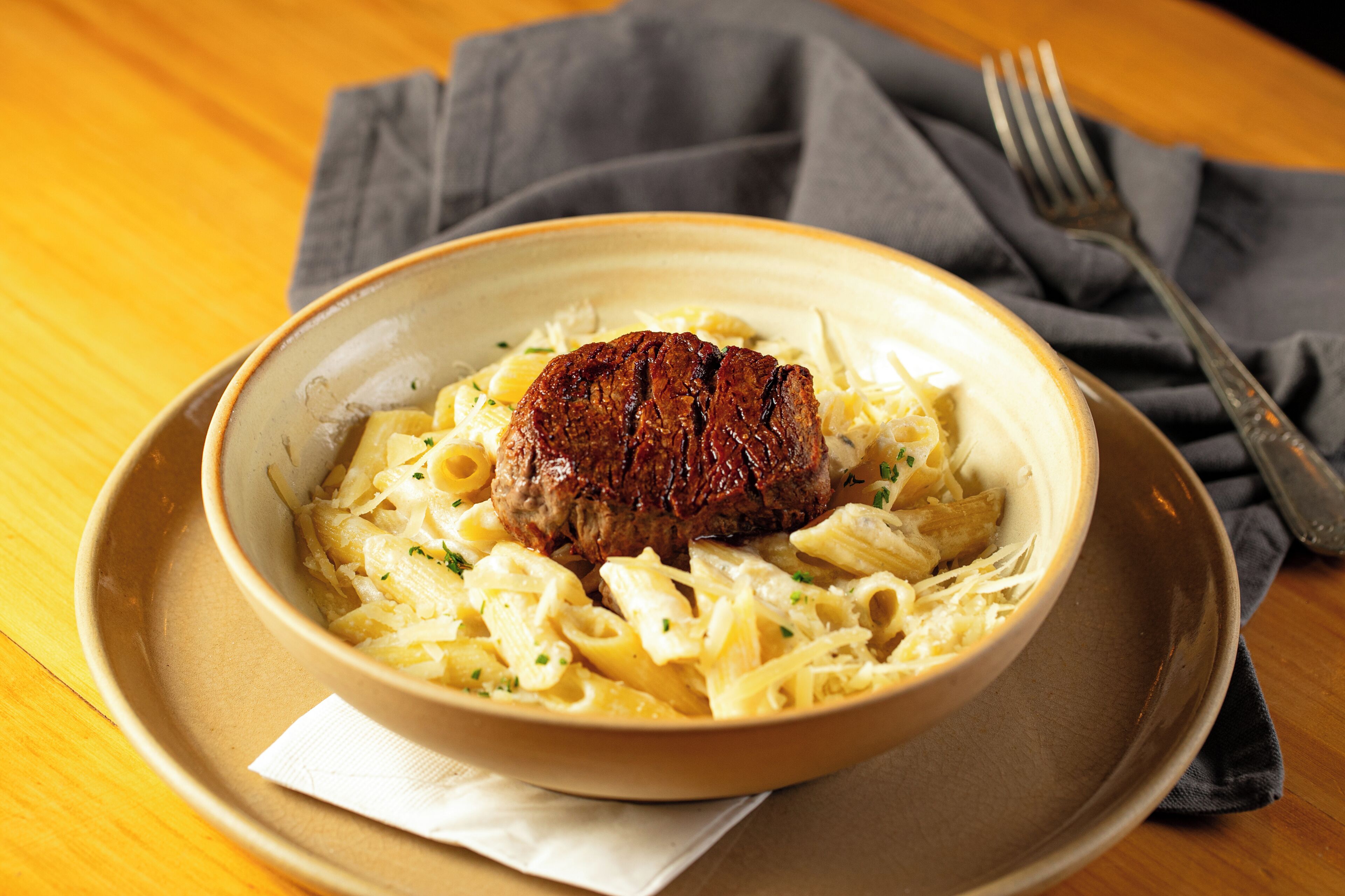 A bowl of pasta with a steak on a plate, placed on a wooden table with a napkin and a fork beside it.