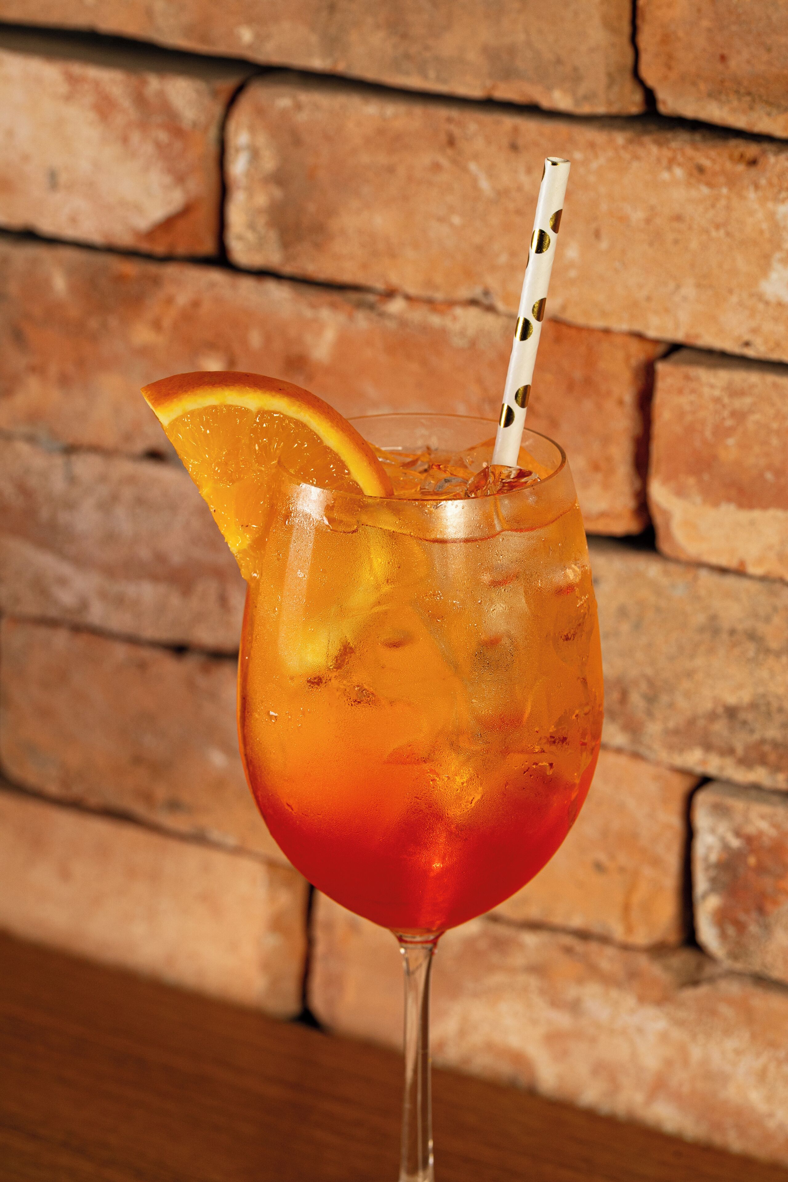Un vaso con una rodaja de naranja y una pajita, sobre una superficie de madera con un muro de ladrillo al fondo.