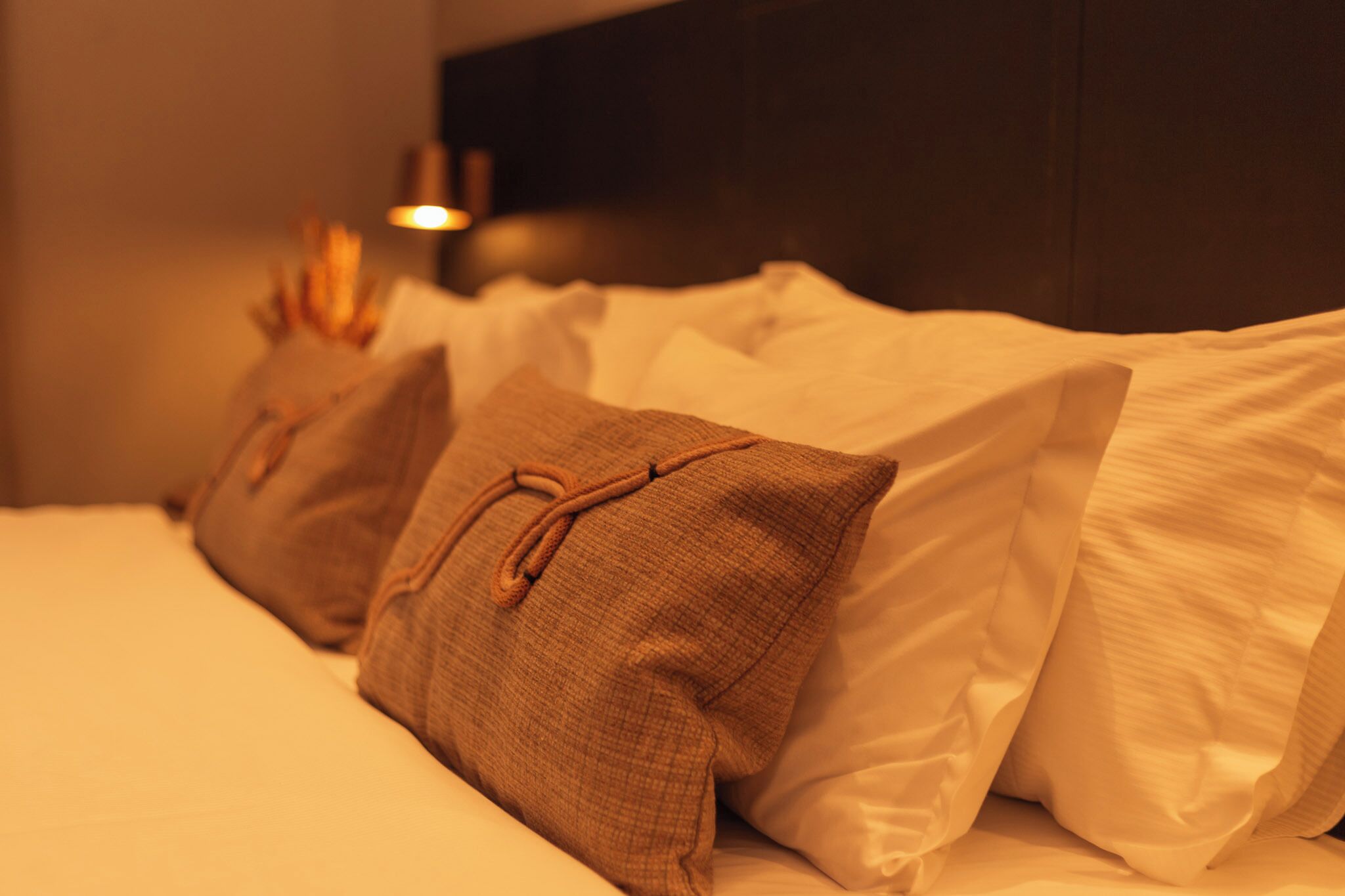 Una cama bien tendida con almohadas blancas y marrones, un cabecero oscuro y una mesita de noche con una lámpara.
