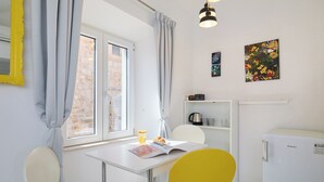 A small, bright room with a white table, two chairs, a window with curtains, a refrigerator, and a shelf with kitchen items. - Room Nino 2 - One Bedroom Apartment, Sleeps 2 (Dubrovnik)