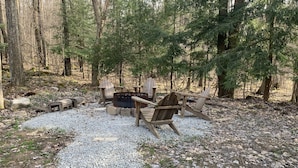 A forested campsite with a fire pit, wooden chairs, and a small table. - Cabin w/ Hot Tub on Small Pond (Rockwood)