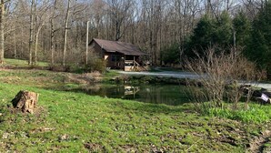 A rustic cabin with a porch, surrounded by a pond and dense trees. - Cabin w/ Hot Tub on Small Pond (Rockwood)