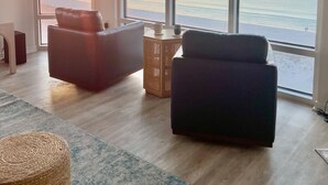Una sala de estar moderna con un ventanal grande que da al océano al atardecer, con dos sofás y una pequeña mesa de madera.