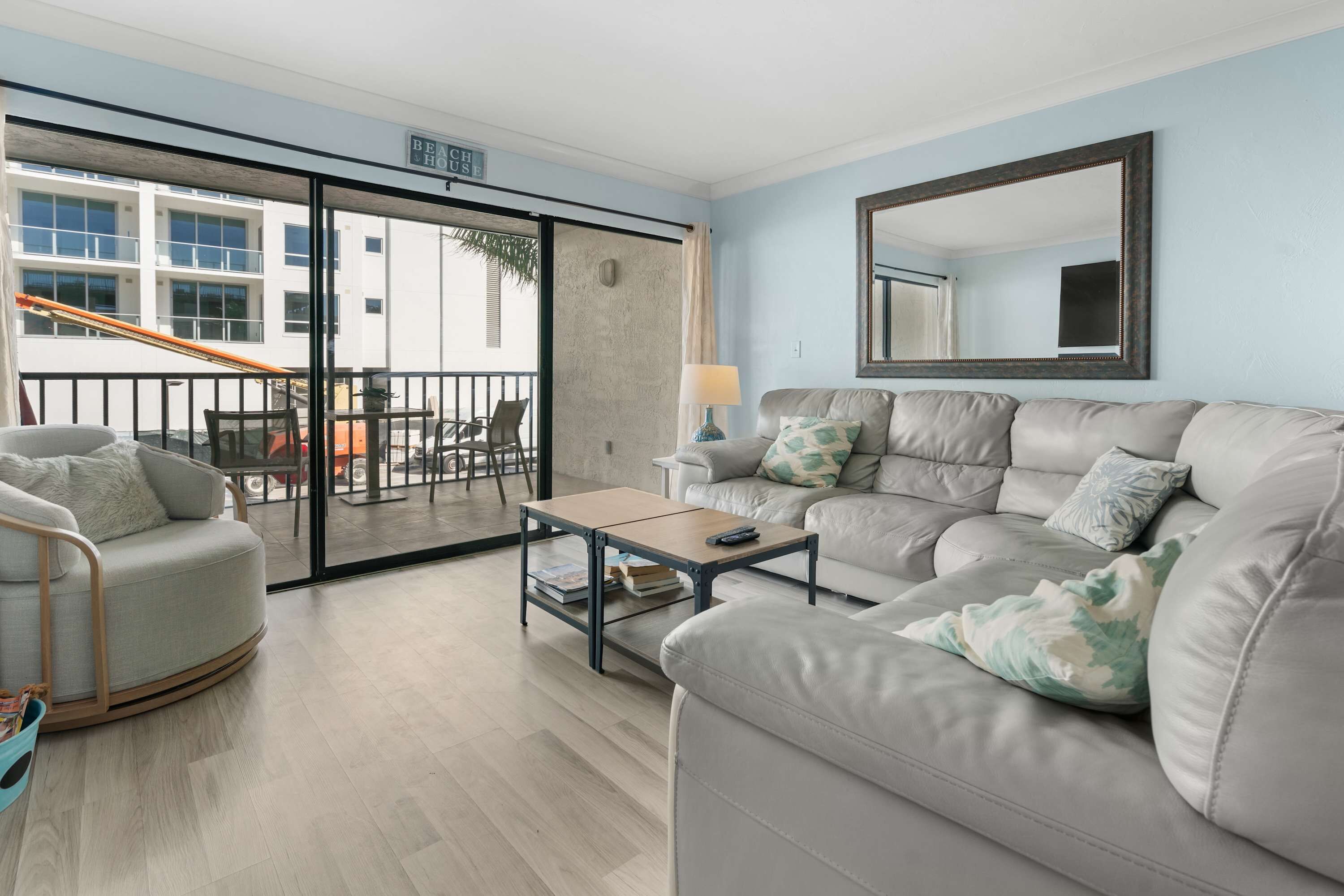 Un salon spacieux avec un canapé gris, une petite table basse et un grand miroir. Une porte-fenêtre coulissante donne accès à un espace extérieur doté d’un balcon.