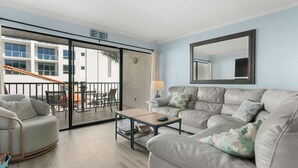 A spacious living room with a grey sofa, a small coffee table, and a large mirror. A sliding glass door leads to an outdoor area with a balcony. - Cozy Condo With Pool Steps From Vanderbilt Beach (Naples)