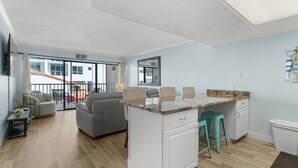 A modern kitchen with a central island, a dining area with a sofa, and a view of the outside. - Cozy Condo With Pool Steps From Vanderbilt Beach (Naples)