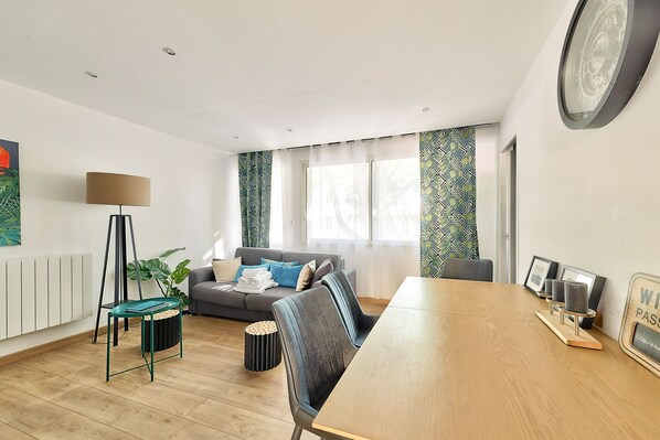 A modern living room with a wooden dining table, a grey sofa, a floor lamp, and a large window with patterned curtains.