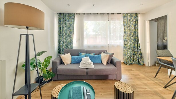 Een moderne woonkamer met een grijze bank, een vloerlamp, een eettafel en een potplant.