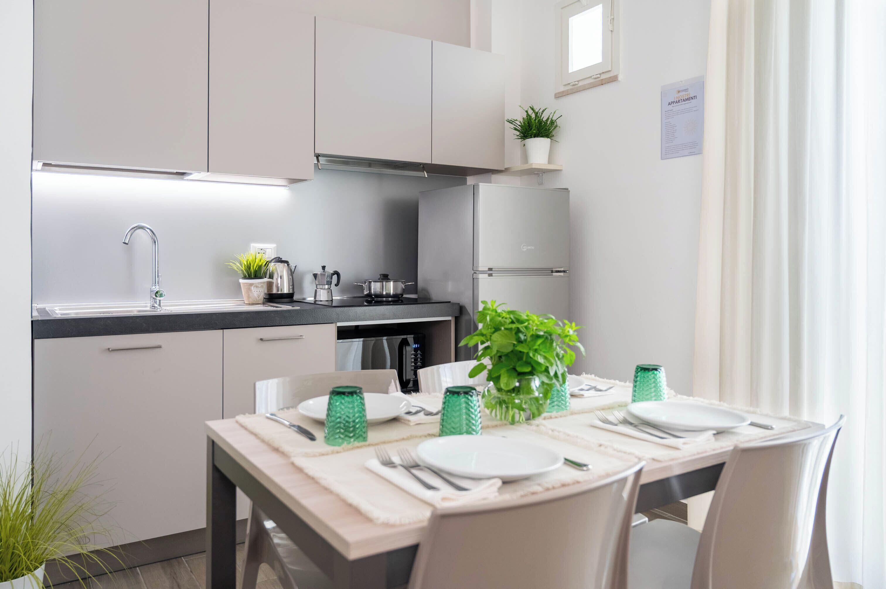 A modern kitchen with white cabinets, a stainless steel refrigerator, and a dining area set with a table, chairs, and green glassware.