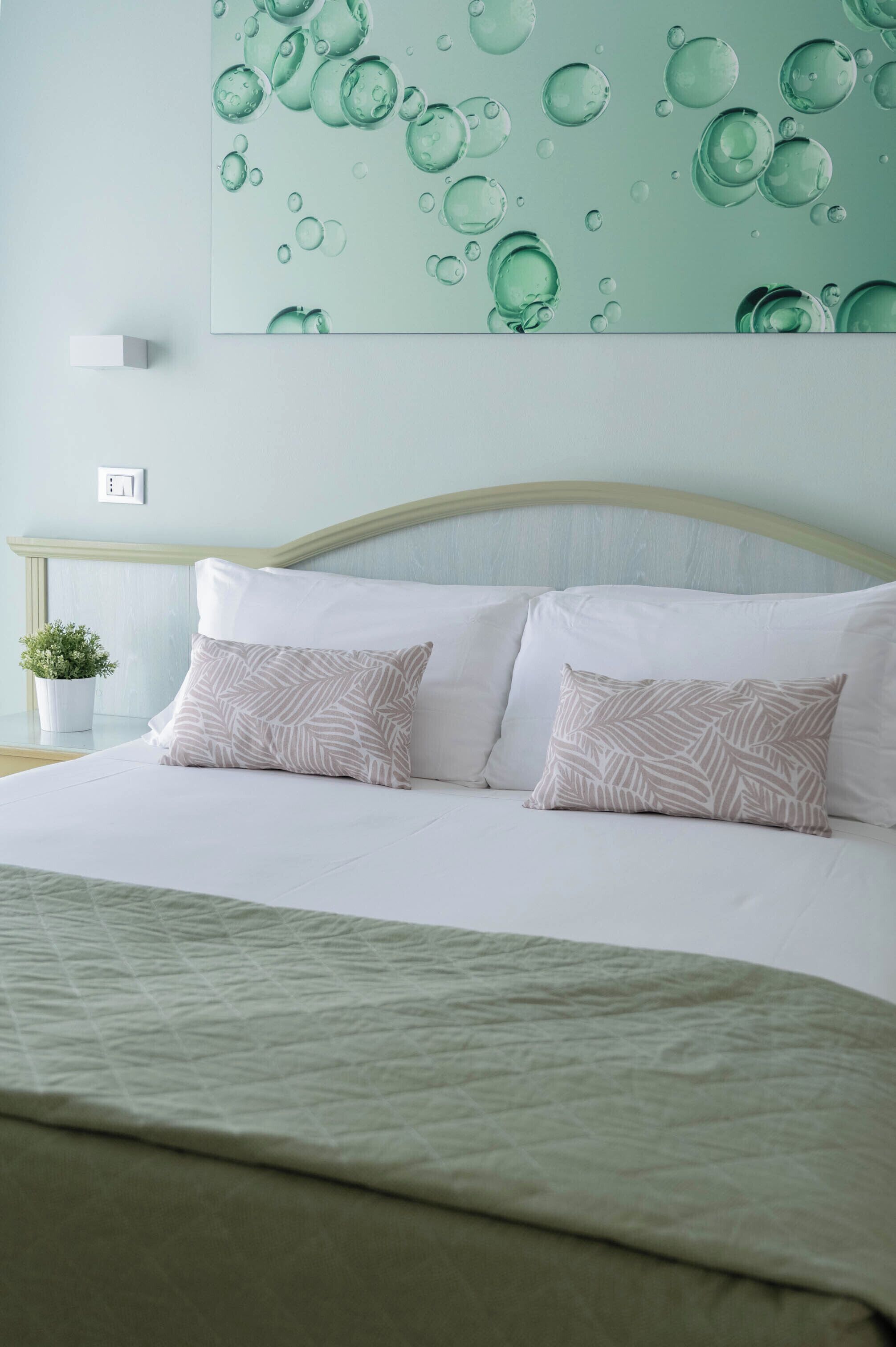 Ein ordentlich bezogenes Bett mit zwei gemusterten Kissen, einer grünen Steude und einer Wand mit Wabenmuster.