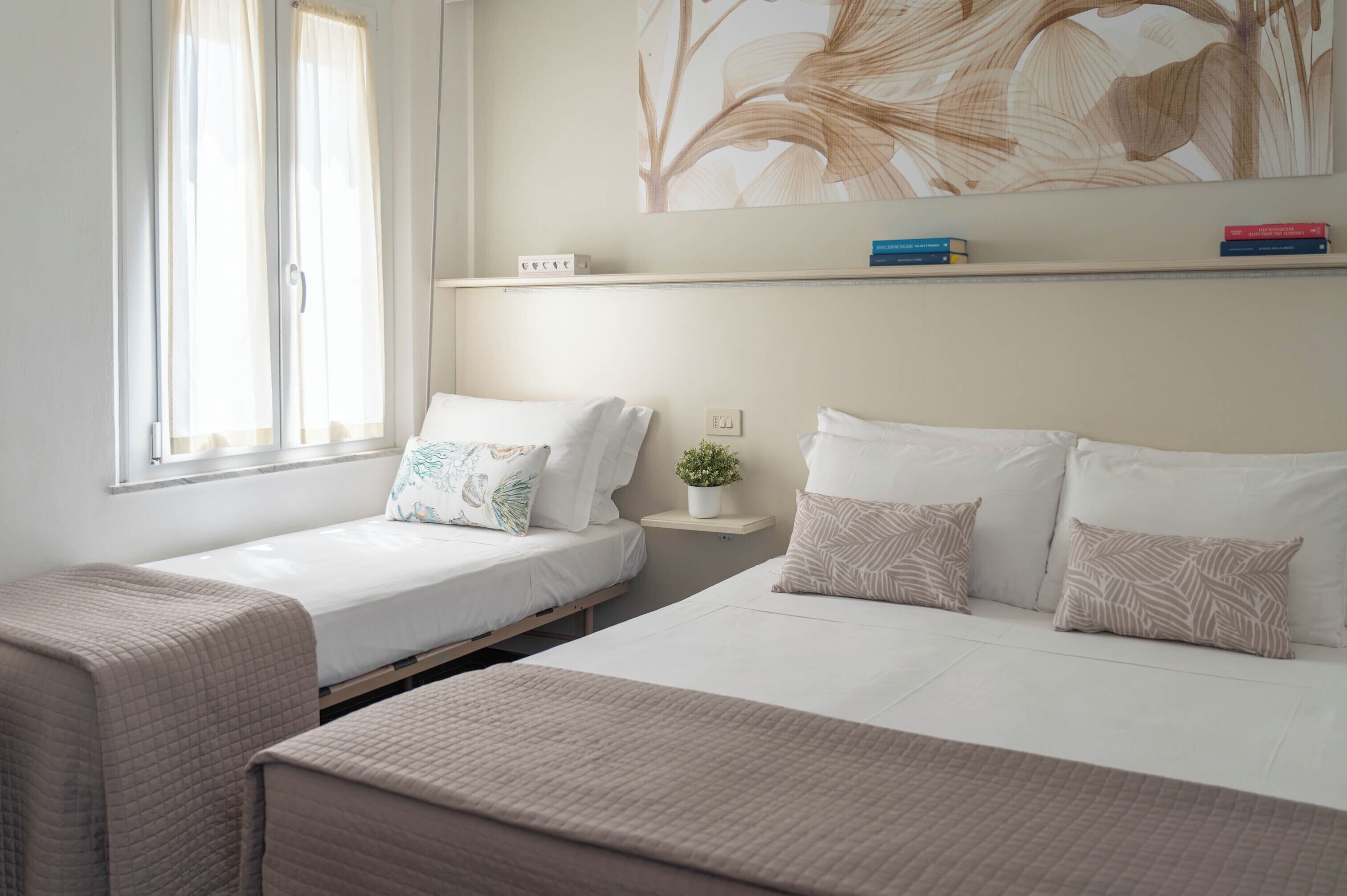 A bedroom with two single beds, a bedside table with a plant, and a shelf with books.