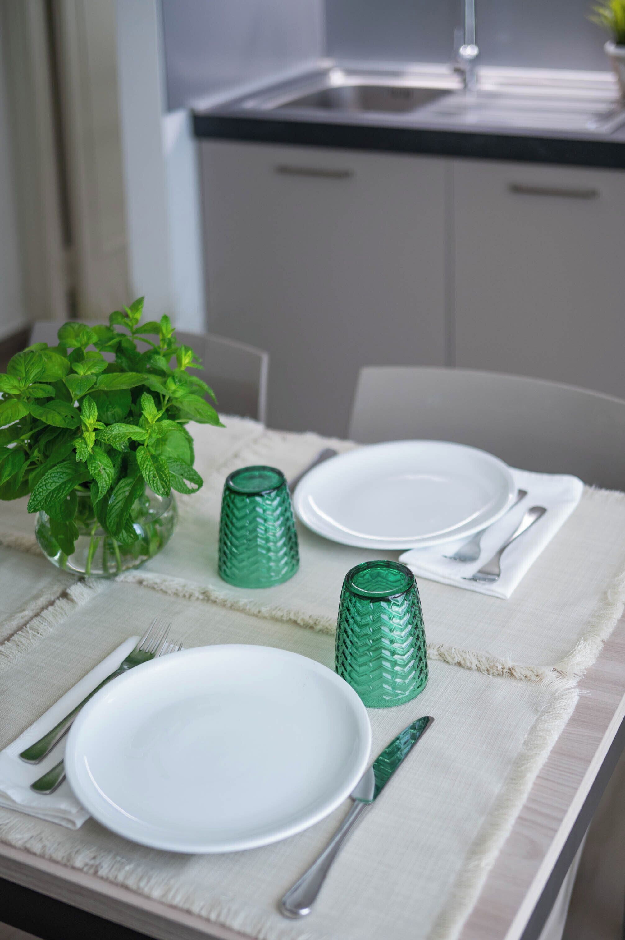 Una mesa de comedor dispuesta con platos blancos, saleros y pimenteros de vidrio verde, y una planta verde en un florero de vidrio transparente.