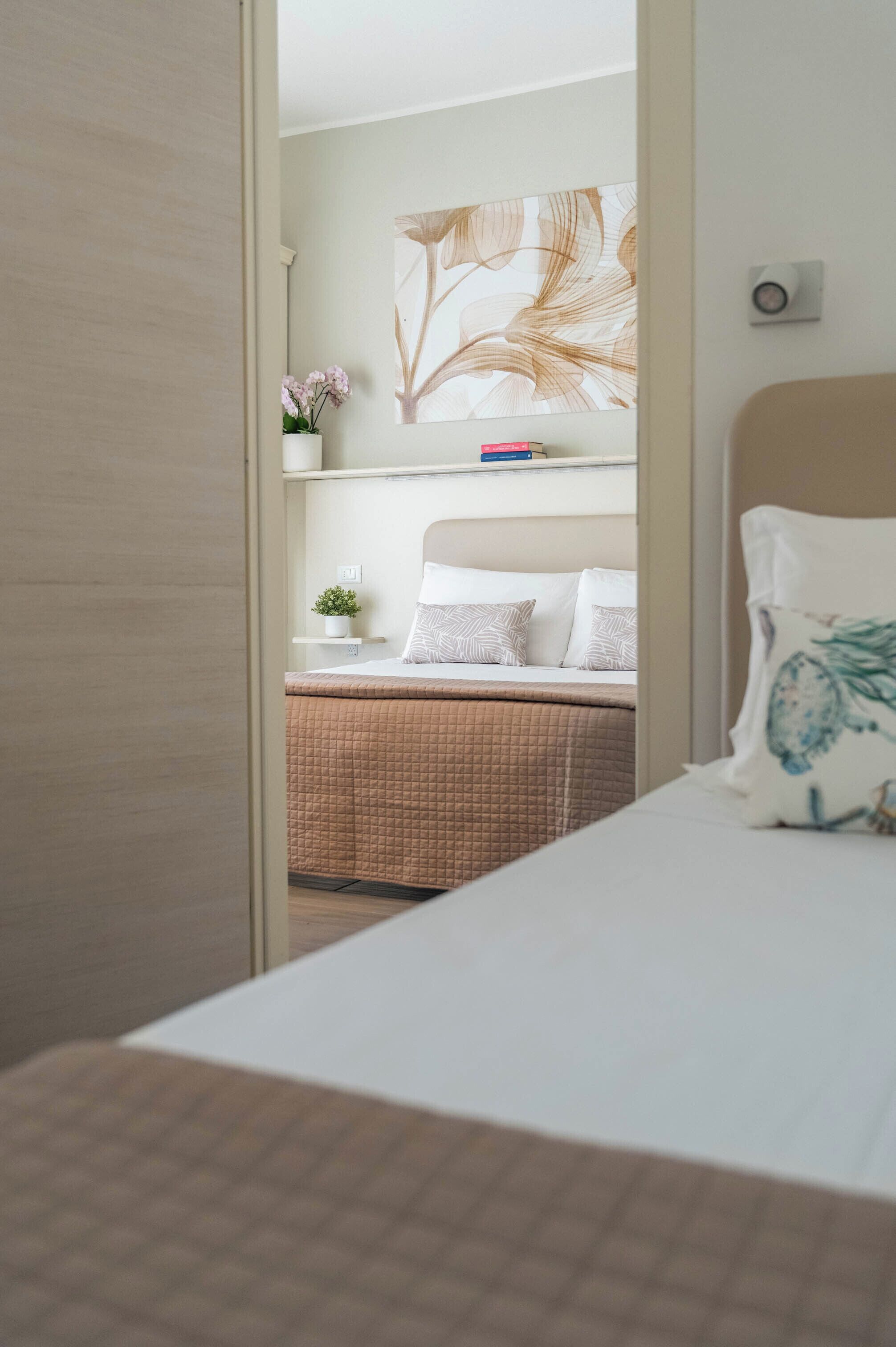 A bedroom with a bed, a bedside table, a nightstand, a wall-mounted shelf with a plant, and a large abstract painting.