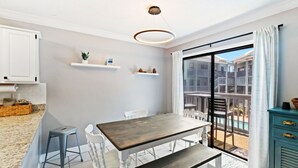 Una cocina moderna con armarios blancos, una mesa de madera y un banco. Hay sillas blancas y un taburete pequeño. Se observa una lámpara en el techo.