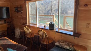 Ein Interieur einer Holzhütte mit einem großen Fenster, das auf eine Terrasse und Bäume blickt. Zwei Holzstühle, ein kleiner Tisch und ein an der Wand befestigtes Fernsehgerät sind vorhanden.