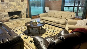 A cozy living room with a stone fireplace, leather sofas, and a large window offering a view of the outdoors. - Rural western cabin- HOT TUB-POOL just 5 mins from exciting Stephenville! (Stephenville)