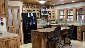 A kitchen with wooden cabinets, a black refrigerator, a microwave, and a dining area with chairs. - Rural western cabin- HOT TUB-POOL just 5 mins from exciting Stephenville! (Stephenville)