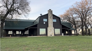 A large, two-story log cabin with a stone chimney, surrounded by a grassy yard and trees. - Rural western cabin- HOT TUB-POOL just 5 mins from exciting Stephenville! (Stephenville)