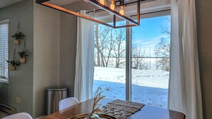Un'area pranzo con un tavolo in legno, un vaso di vetro con piante secche e vista su un paesaggio innevato attraverso ampie finestre.