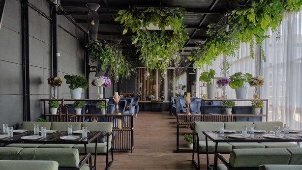 A restaurant with tables set for dining, surrounded by green upholstered chairs, and decorated with hanging plants. - Panarams Tashkent Hotel, A member of Radisson Individuals (Tashkent)