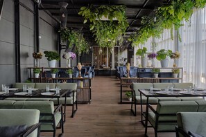 A restaurant with tables set for dining, surrounded by green upholstered chairs, and decorated with hanging plants. - Panarams Tashkent Hotel, A member of Radisson Individuals (Tashkent)