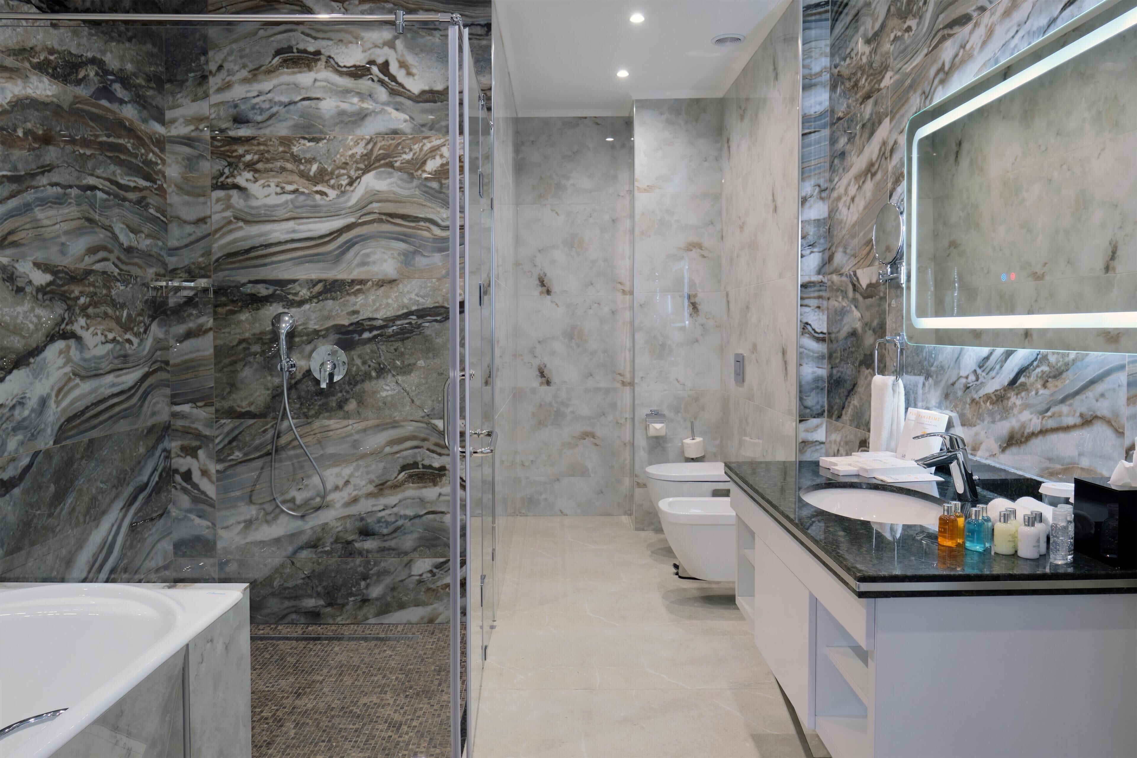 A modern bathroom with a glass shower enclosure, a marble-tiled wall, a large mirror, and a sink with toiletries.