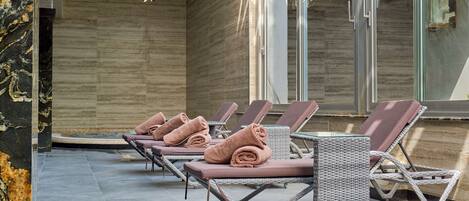 A row of sun loungers with towels, a wicker cart, and a stone wall.