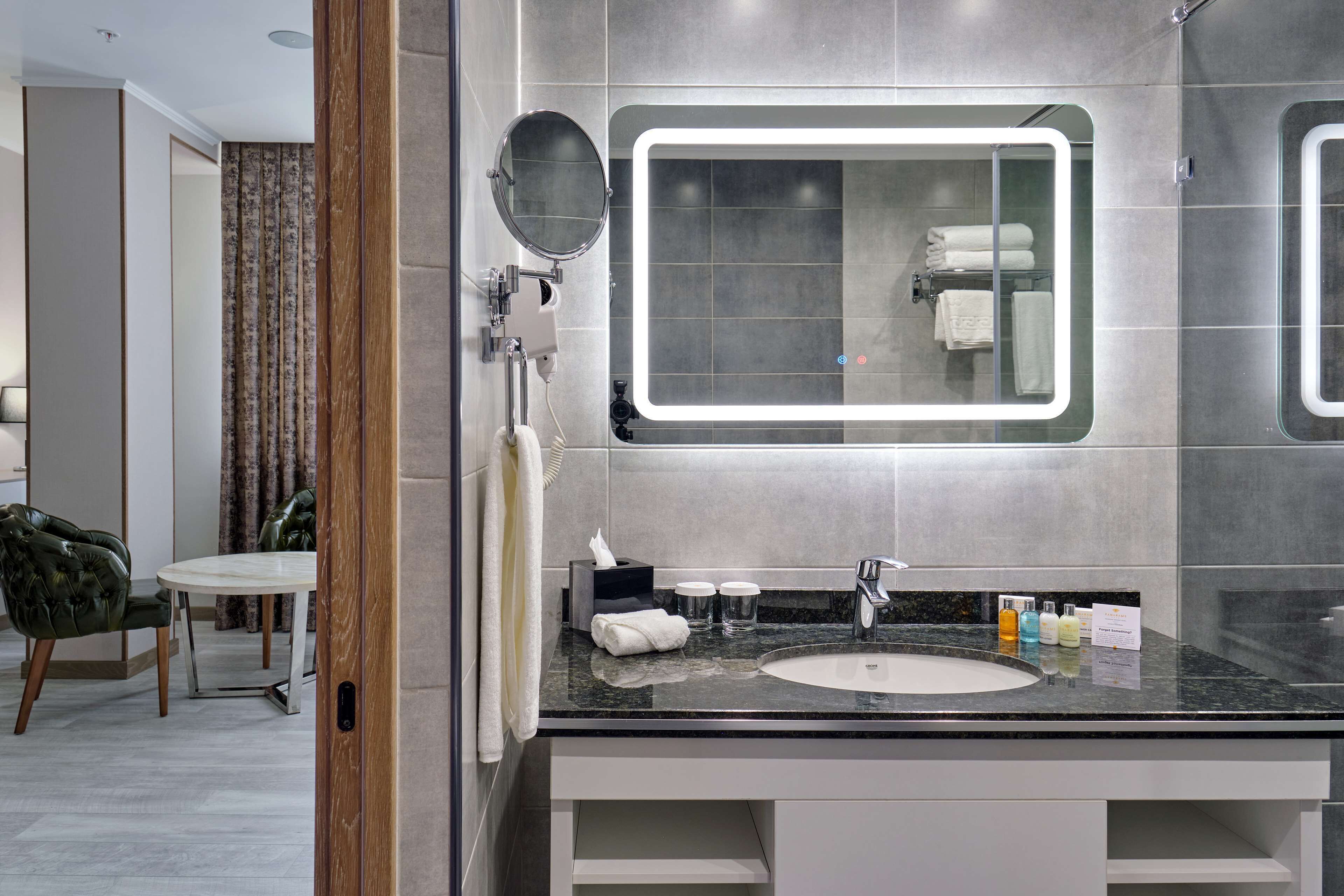 A modern bathroom with a large mirror, a sink, and a toilet. There is a chair and a table in the adjacent room.