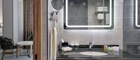 A modern bathroom with a large mirror, a sink, and a toilet. There is a chair and a table in the adjacent room.