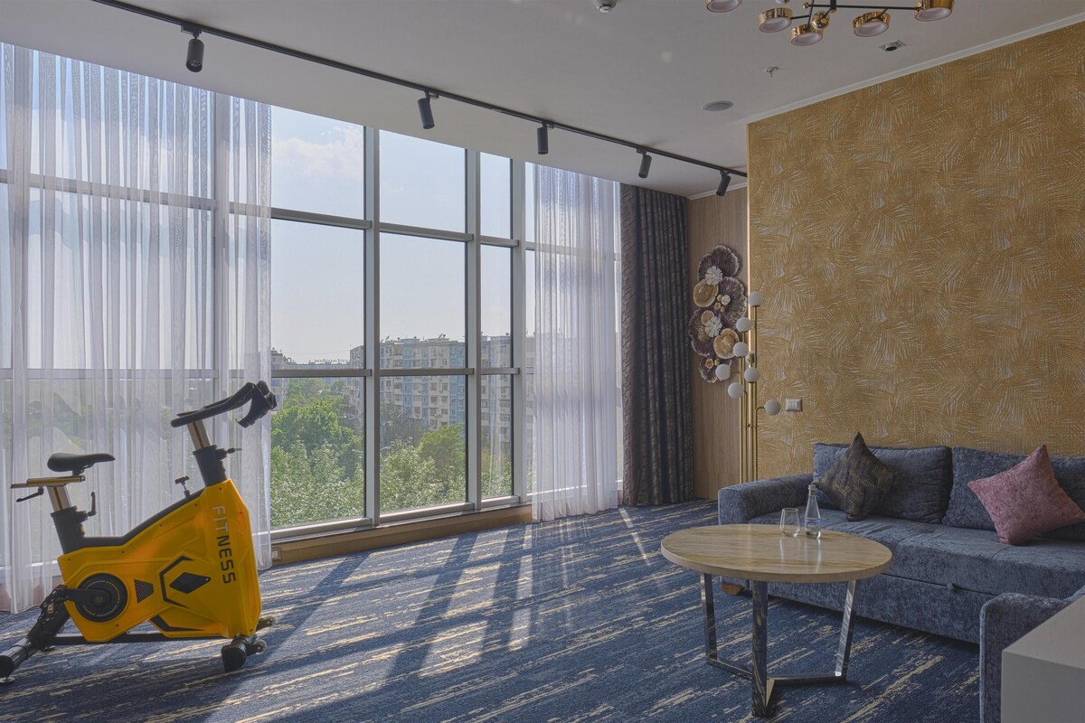 A room with a large window, a sofa, a round table, and an exercise bike.