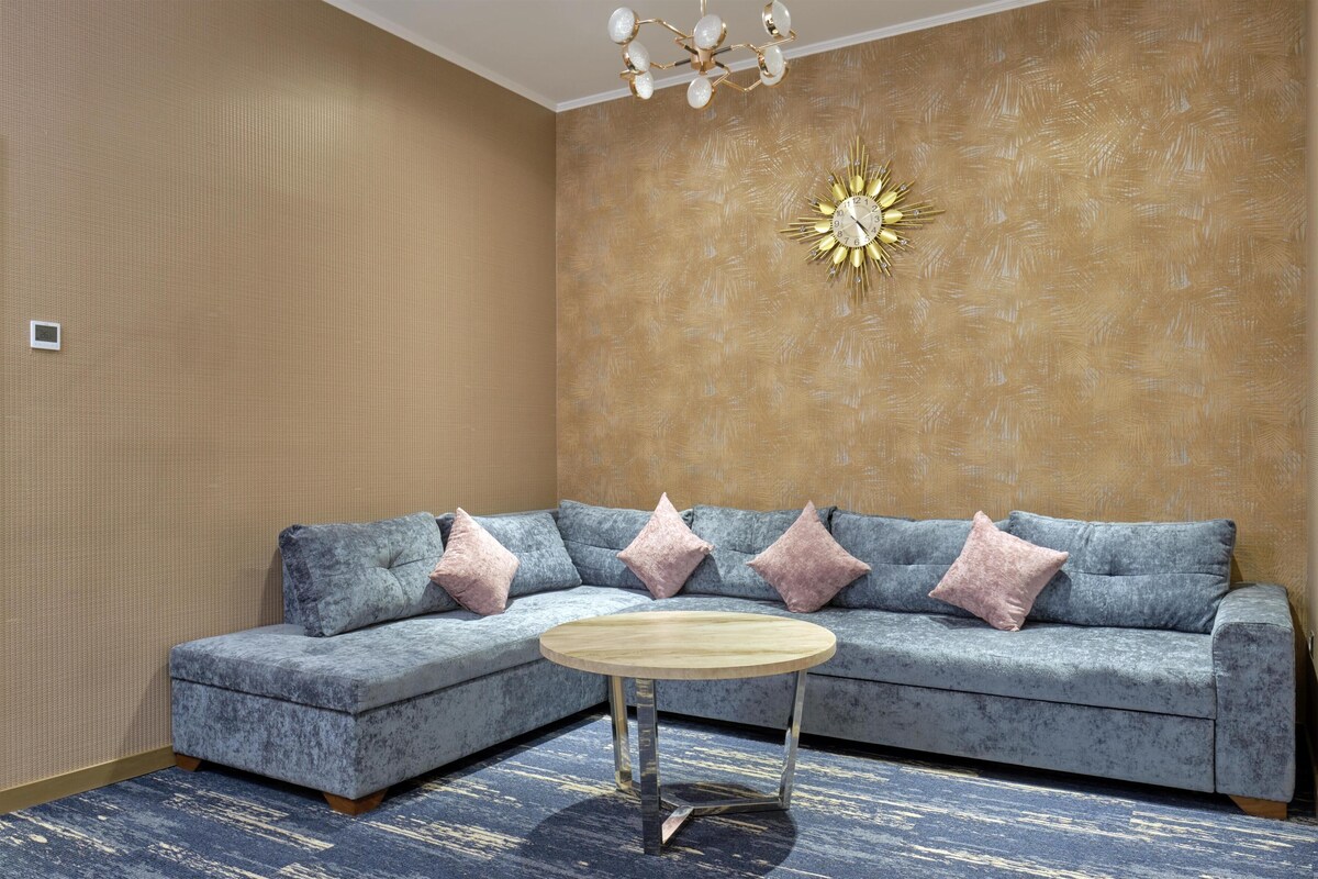 A room with a grey sectional sofa, a round wooden coffee table, and a gold sunburst wall clock.