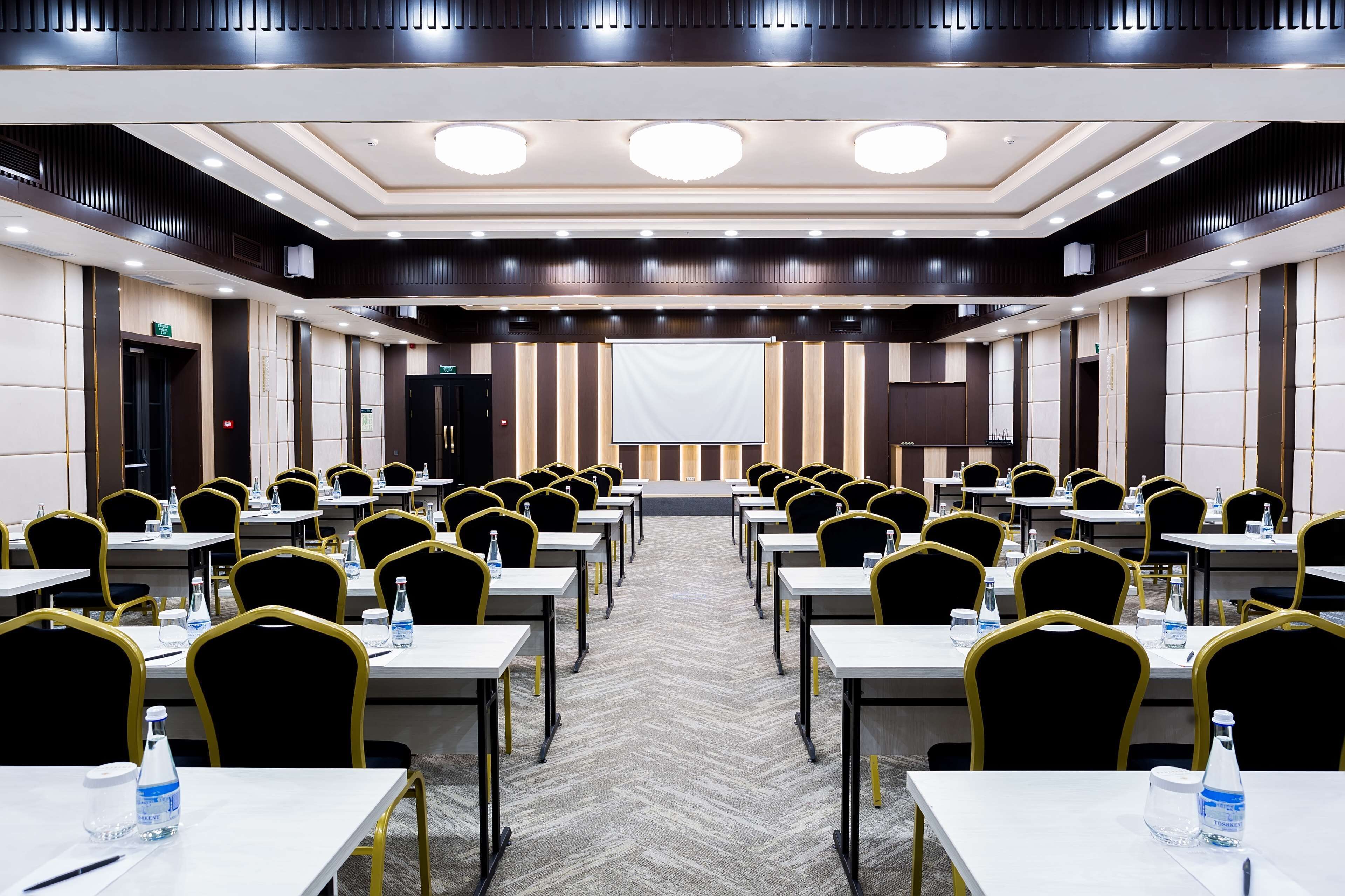 A conference room with rows of tables and chairs, a large screen at the front, and a carpeted floor.