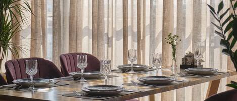 Mesa de jantar posta com pratos, copos e talheres, ladeada por cadeiras, em um quarto com persianas verticais.