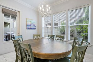 A dining room with a round wooden table, six chairs, a chandelier, and large windows with blinds. - La Terraza 950A - Beach Villa Near the Ocean with Shared Pool (Miramar Beach)