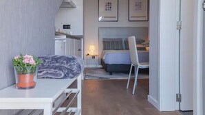 A modern hotel room with a bed, a desk, and a chair. There is a small kitchen area with a refrigerator and a sink. - Infinito Azul II (Caniço)