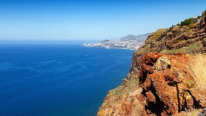 A coastal landscape with a rocky cliff and a view of a distant town across the water. - Infinito Azul II (Caniço)